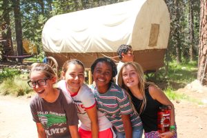 Smiling campers pose at a summer camp in front of a covered wagon