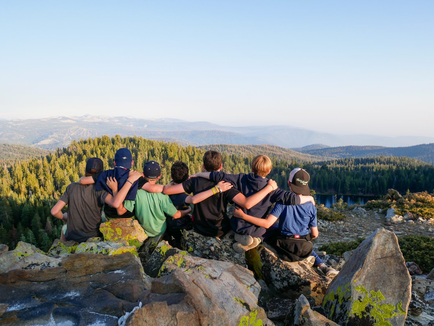 Campers at a summer camp have their arms over each other's shoulders while they look out over the Sierra Nevada and a mountain lake