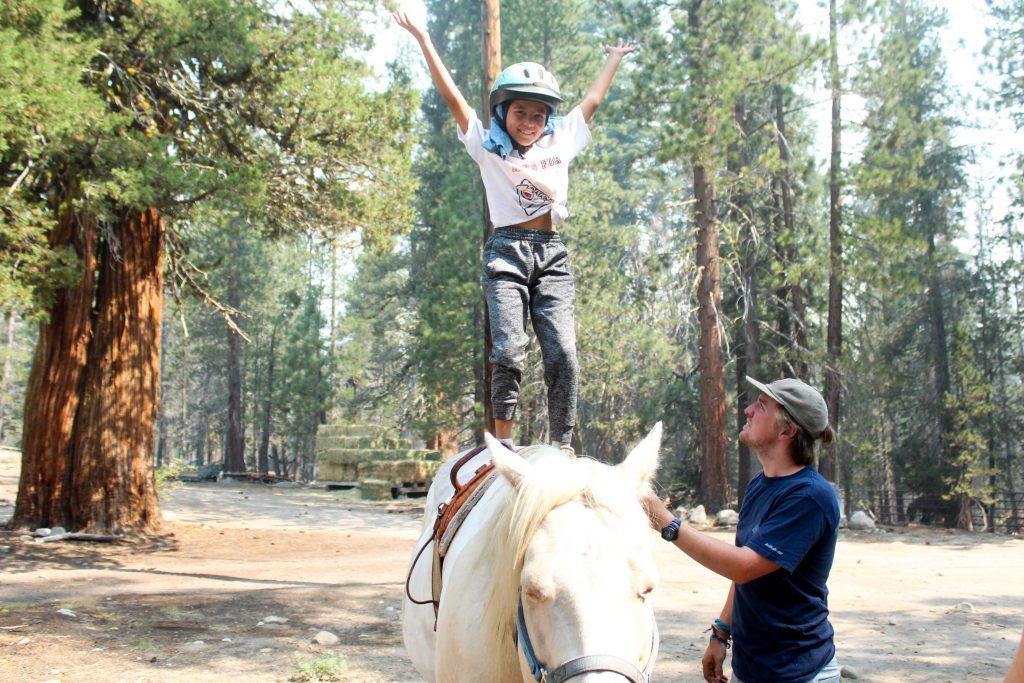 A camper standing on a horse's back with help from a counselor
