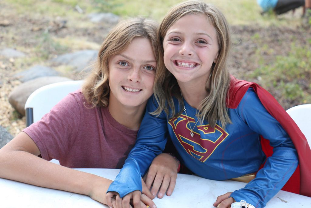 Two summer camp campers smiling at a table, one wearing a super girl costume.